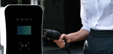 Focus EV charger plug and electric car at public charging station with blur progressive businesswoman holding charger and apartment condo building in background. Eco friendly electric vehicle concept.の写真素材