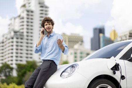 Progressive businessman talking on the phone, leaning on electric car recharging with public EV charging station, apartment condo residential building on the background as green city lifestyle.の写真素材