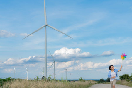 Progressive young asian boy playing with wind pinwheel toy in the wind turbine farm, green field over the hill. Green energy from renewable electric wind generator. Windmill in the countryside conceptの写真素材