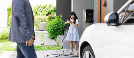 Progressive father and daughter plugs EV charger from home charging station to electric vehicle. Future eco-friendly car with EV cars powered by renewable source of clean energy.の写真素材