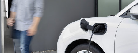 Focus electric car charging at home charging station with blurred progressive man walking in the background. Electric car using renewable clean for eco-friendly concept.の写真素材