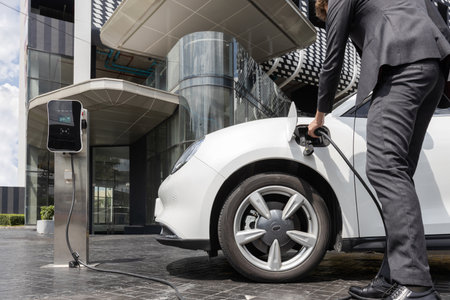 Below view closeup image of progressive black suit businessman recharge battery of his electric vehicle from public charging station. Renewable and alternative energy powered car concept.の写真素材