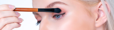 Closeup beautiful personable girl with flawless applying eye shadow makeup with eyeliner brush. Cosmetic facial painting process on lovely young woman with perfect clean skin in isolated background.の写真素材