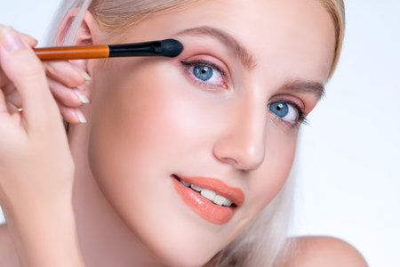 Closeup beautiful personable girl with flawless applying eye shadow makeup with eyeliner brush. Cosmetic facial painting process on lovely young woman with perfect clean skin in isolated background.の写真素材