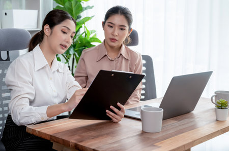 Two young office lady colleagues collaborating in modern office workspace, engaging in discussion and working together on laptop, showcasing their professionalism as modern office worker. Enthusiasticの写真素材