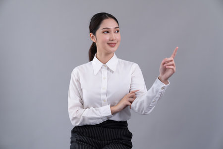 Asian female call center operator with smile face advertises job opportunity, wearing a formal suit and headset pointing finger for product on customizable isolated background. Enthusiasticの写真素材