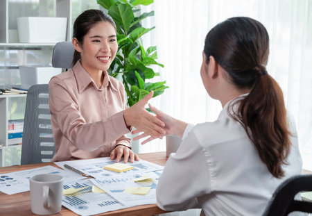 Two Asian businesswomen shaking hands in modern office after successfully analyzing piles of dashboard data papers, as importance of teamwork and integrity in the workplace concept. Enthusiasticの写真素材