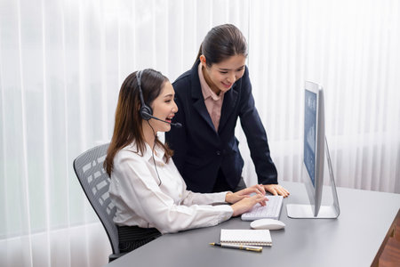Asian female customer support operator wearing headset is guided by her supportive manager. Experienced colleague help operator handle a call with a client, providing advice and guidance. Enthusiasticの写真素材