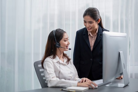 Asian female customer support operator wearing headset is guided by her supportive manager. Experienced colleague help operator handle a call with a client, providing advice and guidance. Enthusiasticの写真素材