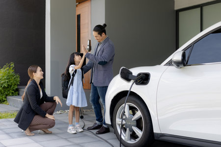 Progressive young parents and daughter with electric vehicle and home charging station. Green and clean energy from electric vehicles for healthy environment. Eco power from renewable source at home.の写真素材