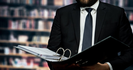 Sharp-suited businessman reviews and works on financial reports and paperwork with focused attention in his office with background of bookshelf in library. Equilibriumの写真素材