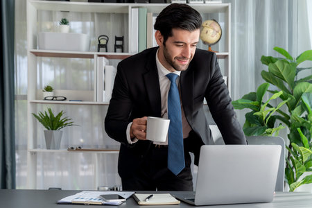 Modern professional businessman at modern office desk using laptop to work with coffee in his hand....