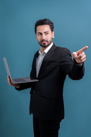 Successful businessman in black suit with innovative tech concept, finger pointing at camera and holding laptop and smiling with excitement on copyspace background for promotion or ad. Ferventの写真素材