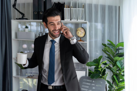 Diligent and busy businessman talking on phone call with coworker in modern office. Making sales call to client with coffee in hand, standing posing confidently as professional office worker. Ferventの写真素材