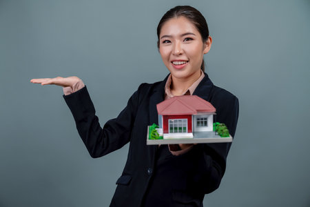 Young Asian woman wearing formal suit, holding and showcasing house model on isolated background. Ideal for housing advertisement, home insurance promotions, or customizable copy space. Enthusiasticの写真素材