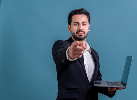 Successful businessman in black suit with innovative tech concept, finger pointing at camera and holding laptop and smiling with excitement on copyspace background for promotion or ad. Ferventの写真素材