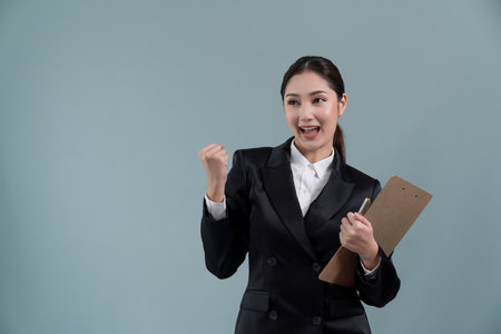 Confident young asian businesswoman in formal suit holding clipboard with hand gesture indicating promotion or advertising with surprised facial expression on isolated background. Enthusiasticの写真素材