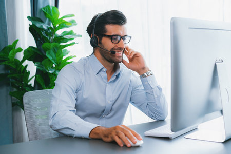 Male call center operator or telesales representative siting at his office desk wearing headset and...