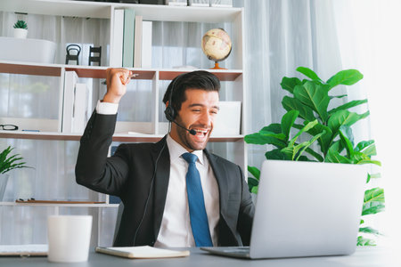 Excited and happy businessman dressed in black formal suit, raise his arm in celebratory gesture at office desk. Successfully call center or telesales operator celebrate in workplace. Ferventの写真素材