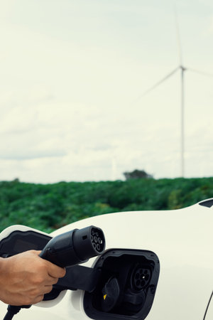 Progressive natural scenic with windmill generator where hand insert charging plug to electric vehicle from charging station with natural background. EV car powered by wind turbine electric generator.の写真素材
