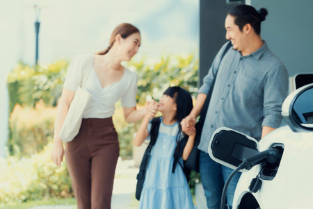 Focus closeup electric vehicle recharging battery from home electric charging station with blurred family in background. Renewable clean energy car for progressive eco awareness lifestyle concept.の写真素材