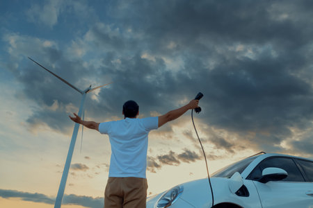 Progressive man spread arm with electric car recharge energy from charging station on green field powered by wind turbine concept of future sustainable energy.の写真素材