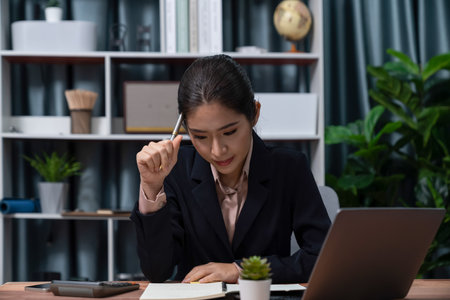 Young asian businesswoman is diligently working and taking notes in her modern office workspace, demonstrating professionalism as attractive and diligent office lady. Enthusiasticの写真素材