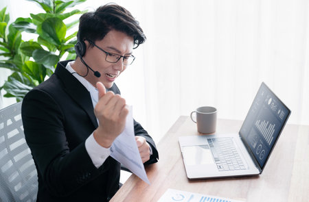 Confident asian telesales businessman work in modern office wearing headphone, presenting BI dashboard analysis to clients and customers in online meeting. Success modern telesales business . Jubilantの写真素材