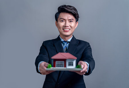 Confident Asian businessman holding house model, advertising home loan with smile. Real estate agent with sample house model in hand on isolated background for housing business advertisement. Jubilantの写真素材
