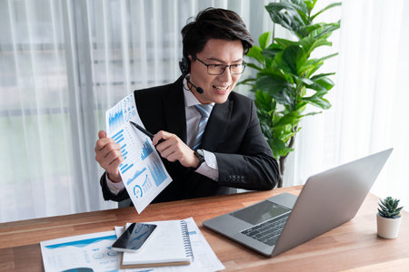 Confident asian telesales businessman work in modern office wearing headphone, presenting BI dashboard analysis to clients and customers in online meeting. Success modern telesales business . Jubilantの写真素材
