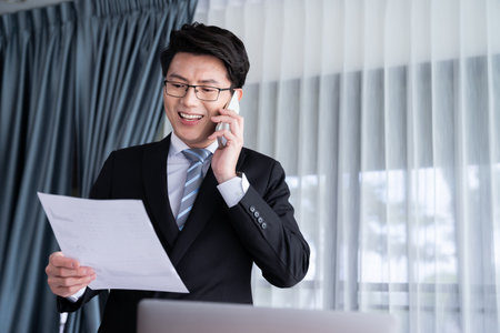 Businessman working on laptop for on office desk workspace with his smartphone. Smart executive researching financial business data or planning strategic business, talking on phone. Jubilantの写真素材