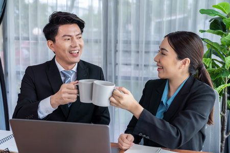 Two Asian office workers taking coffee break together in workplace. Coworkers smiling and socializing while holding cup of coffee adding friendly working environment in corporate workspace. Jubilantの写真素材