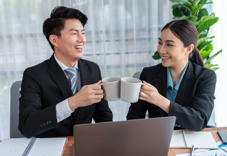 Two Asian office workers taking coffee break together in workplace. Coworkers smiling and socializing while holding cup of coffee adding friendly working environment in corporate workspace. Jubilantの写真素材