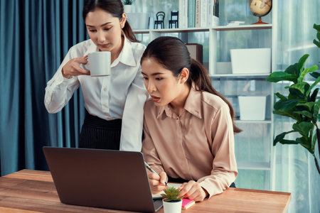 Two young office lady colleagues collaborating in modern office workspace, engaging in discussion and working together on laptop, showcasing their professionalism as modern office worker. Enthusiasticの写真素材