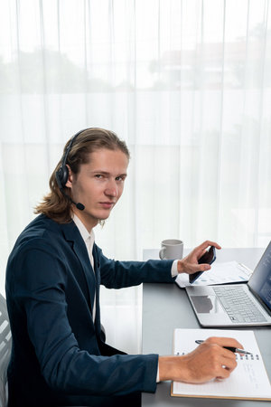 Male customer service operator or telesales agent sitting at desk in office, wearing headset and...