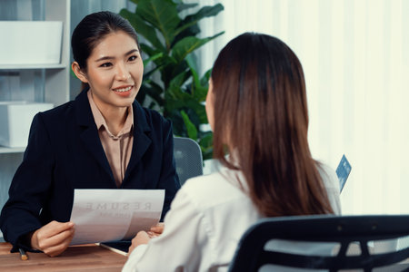 Two asian women conduct job interview in office. Applicants wear formal suit while talking about her CV and job application. Interviewer ask inquiry in positive and conversational manner. Enthusiasticの写真素材