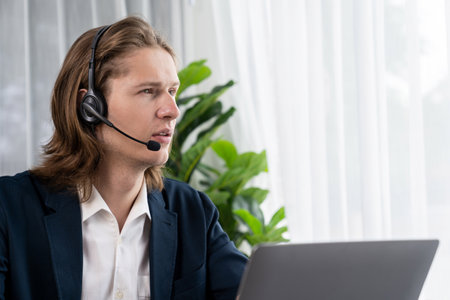 Male customer service operator or telesales agent sitting at desk in office, wearing headset and...