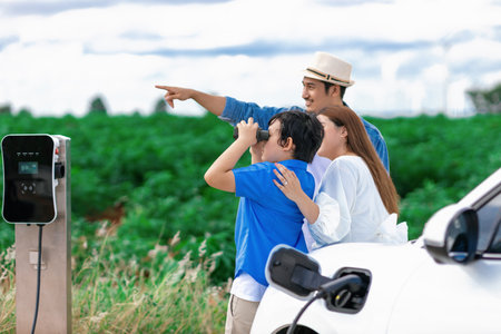 Concept of progressive happy family enjoying their time at wind farm with electric vehicle. Electric vehicle driven by clean renewable energy from wind turbine generator for charging station.の写真素材