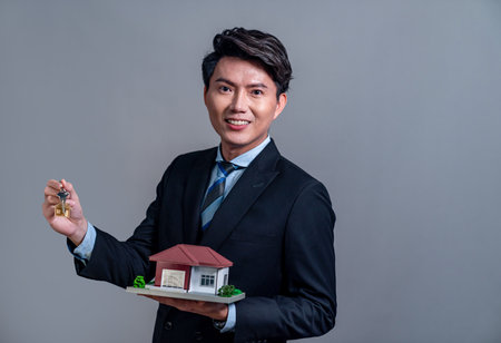 Confident Asian businessman holding house model, advertising home loan with smile. Real estate agent with sample house model in hand on isolated background for housing business advertisement. Jubilantの写真素材