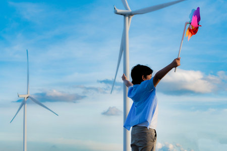 Progressive young asian boy playing with wind pinwheel toy in the wind turbine farm, green field over the hill. Green energy from renewable electric wind generator. Windmill in the countryside conceptの写真素材