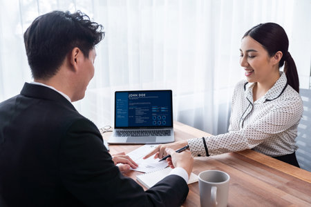 Conversational job interview between interviewer and candidate. Asian female job applicant present her performance and qualification by digital resume on laptop and CV paper to HR manager. Jubilantの写真素材