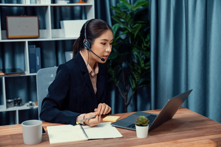 Asian call center with headset and microphone working on her laptop. Female operator provide exceptional customer service. Supportive call center agent helping customer on inquiry. Enthusiasticの写真素材