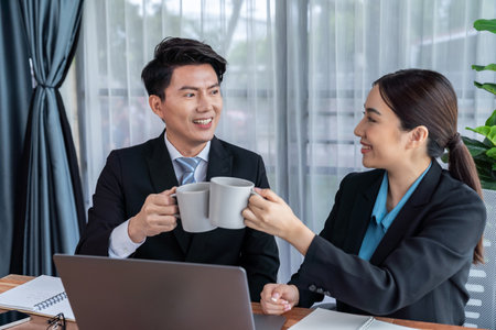 Two Asian office workers taking coffee break together in workplace. Coworkers smiling and socializing while holding cup of coffee adding friendly working environment in corporate workspace. Jubilantの写真素材
