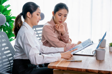 Two young office lady colleagues collaborating in modern office workspace, engaging in discussion and working together on laptop, showcasing their professionalism as modern office worker. Enthusiasticの写真素材