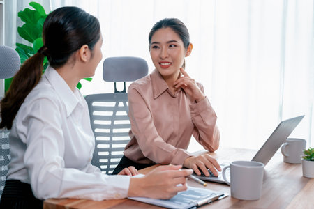 Two young office lady colleagues collaborating in modern office workspace, engaging in discussion and working together on laptop, showcasing their professionalism as modern office worker. Enthusiasticの写真素材