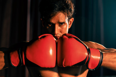 Muay Thai boxer punch fish his hand together in front of camera in ready to fight stance posing at gym with boxing equipment in background. Focused determination eye and prepare for challenge. Impetusの写真素材