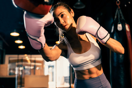 Asian female Muay Thai boxer punching in fierce boxing training session, delivering strike to her sparring trainer wearing punching mitts, showcasing Muay Thai boxing technique and skill. Impetusの写真素材