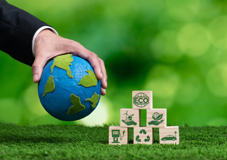 Businessman hand holding wooden cube with eco symbol and paper globe on fertile soil background. Environmental awareness and sustainable energy. Clean and renewable energy for greener ecology. Alterの写真素材