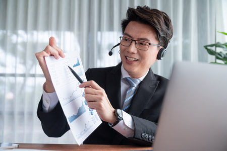 Confident asian telesales businessman work in modern office wearing headphone, presenting BI dashboard analysis to clients and customers in online meeting. Success modern telesales business . Jubilantの写真素材