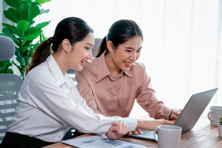 Two young office lady colleagues collaborating in modern office workspace, engaging in discussion and working together on laptop, showcasing their professionalism as modern office worker. Enthusiasticの写真素材